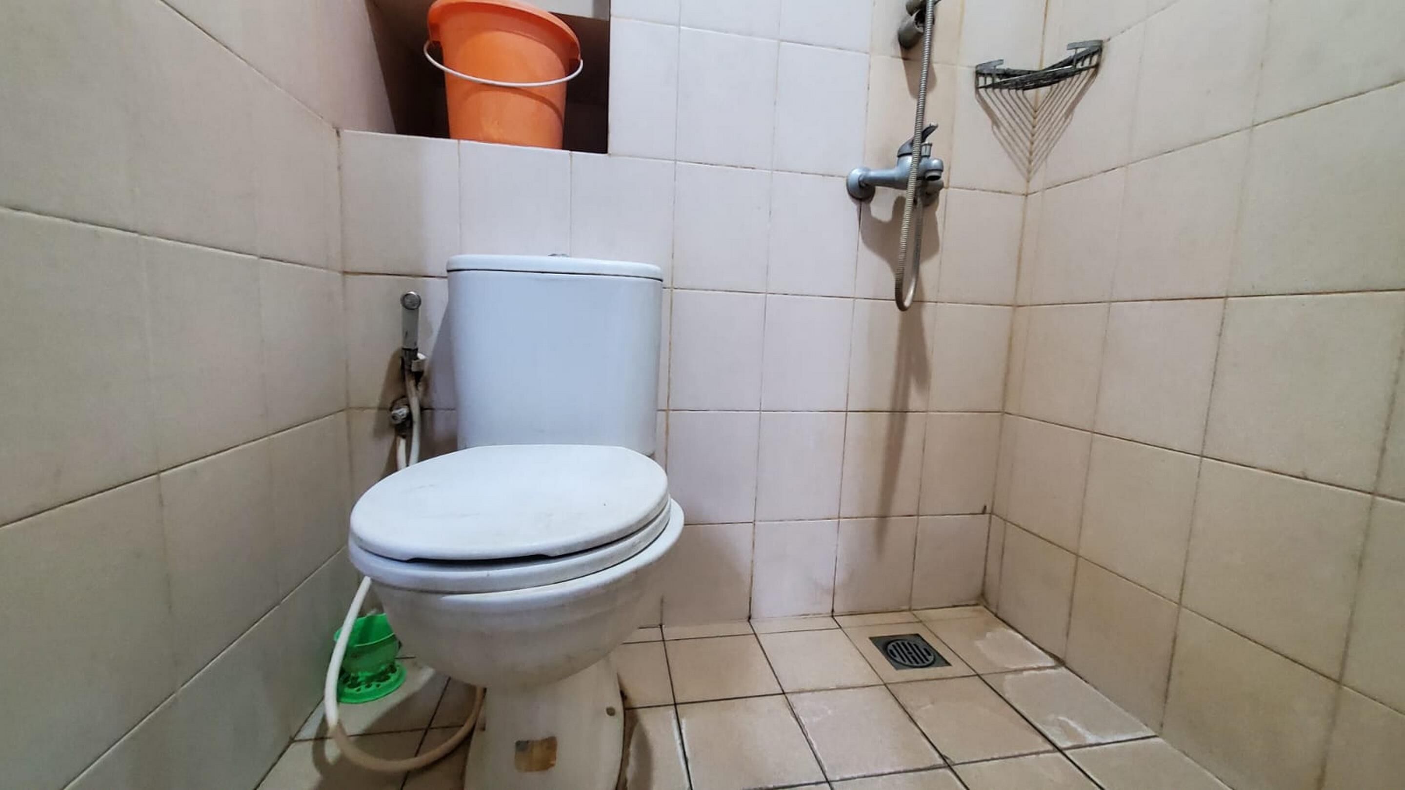 A small, tiled bathroom with a toilet, a shower, and a green cleaning bucket on the wall.