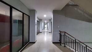 Ein langer, schmaler Korridor mit einer Treppe auf der rechten Seite, die zu einer oberen Etage führt. Die Wände sind schlicht und in einer hellen Farbe gehalten, der Boden ist mit Fliesen belegt.