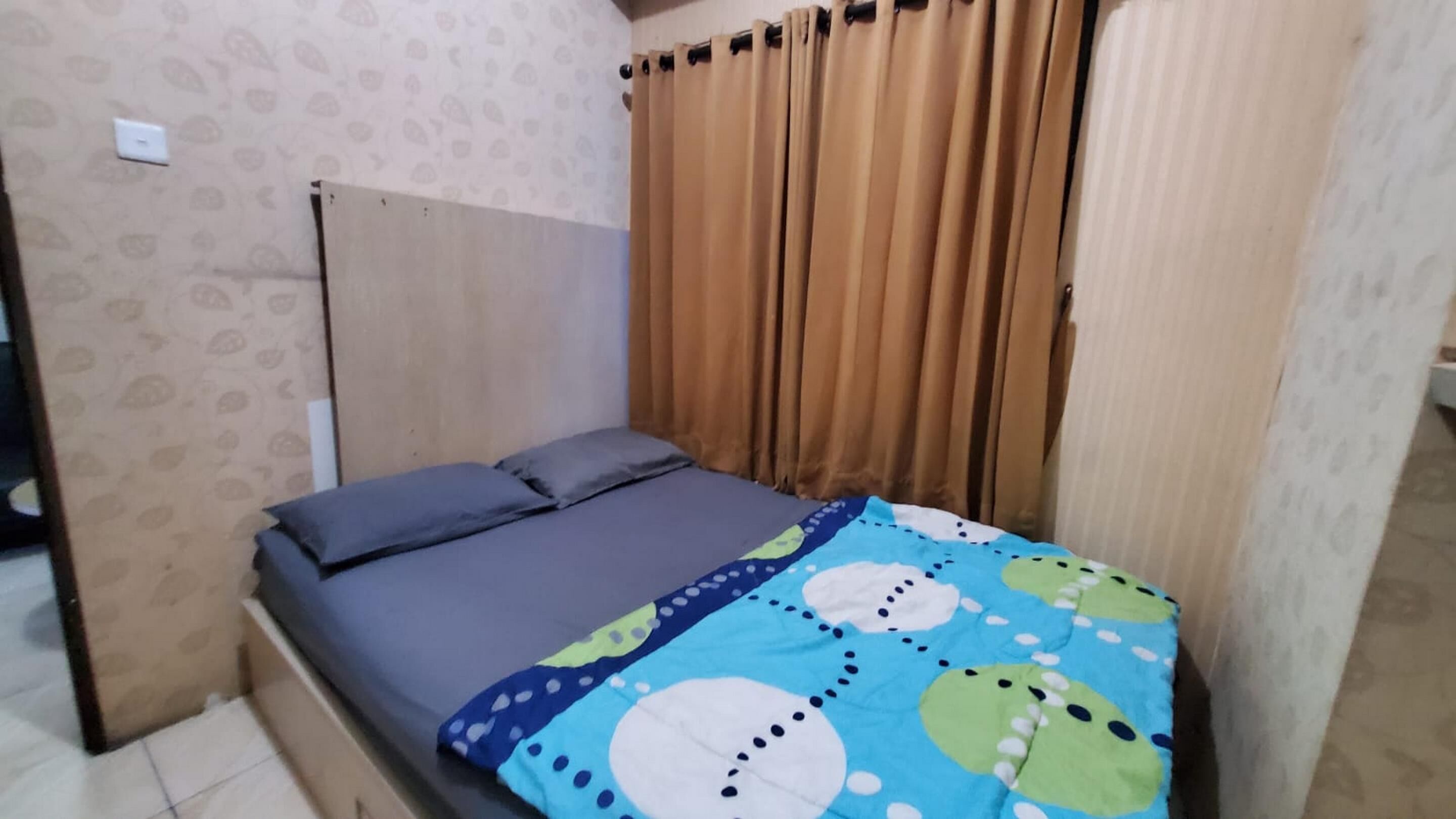 A small, single-bed room with a plain headboard, a patterned wall, and a window with brown curtains.