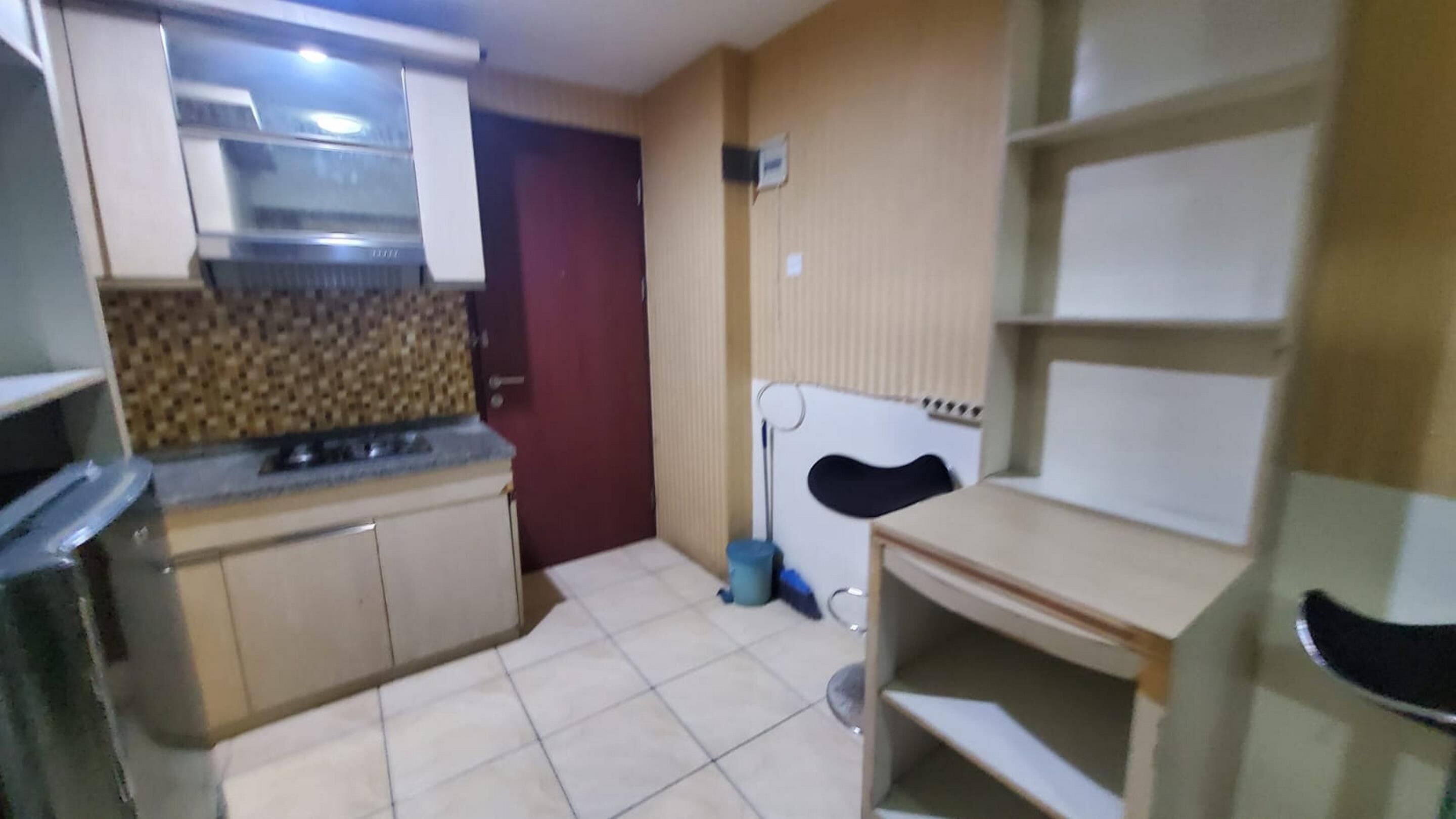Een kleine keuken met een fornuis, kastjes en een magnetron. Er staat een tafeltje met een stoel.