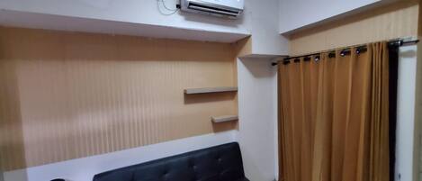 A room with a black sofa, a wall-mounted air conditioner, and a window with brown curtains.