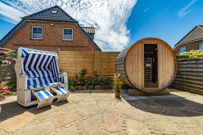 Een stenen terras met een sauna, een houten vat-sauna en een gestreept lounge stoel.