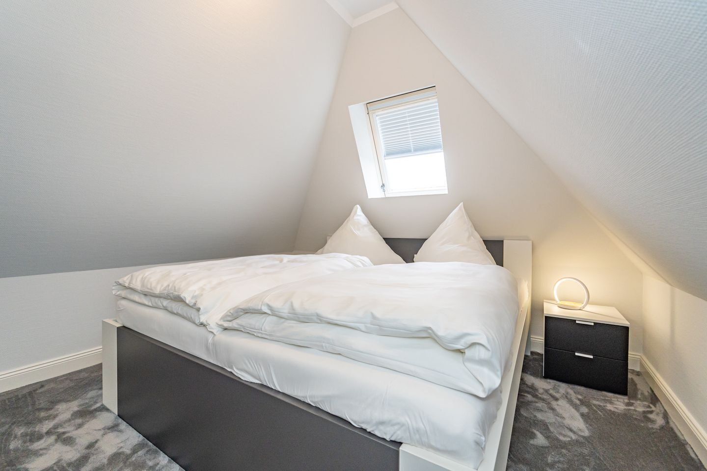 Un dormitorio con cama, mesita de noche, lámpara y ventana.