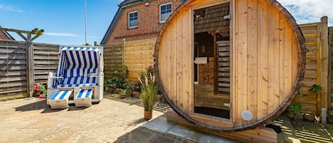 A wooden sauna with a door, a striped lounge chair, and a brick house in the background.