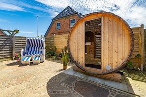 A wooden sauna with a door, a striped lounge chair, and a brick house in the background.