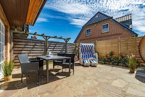 Une terrasse extérieure avec un sol en pierre, une table entourée de chaises, un espace salon abrité et une clôture en bois.