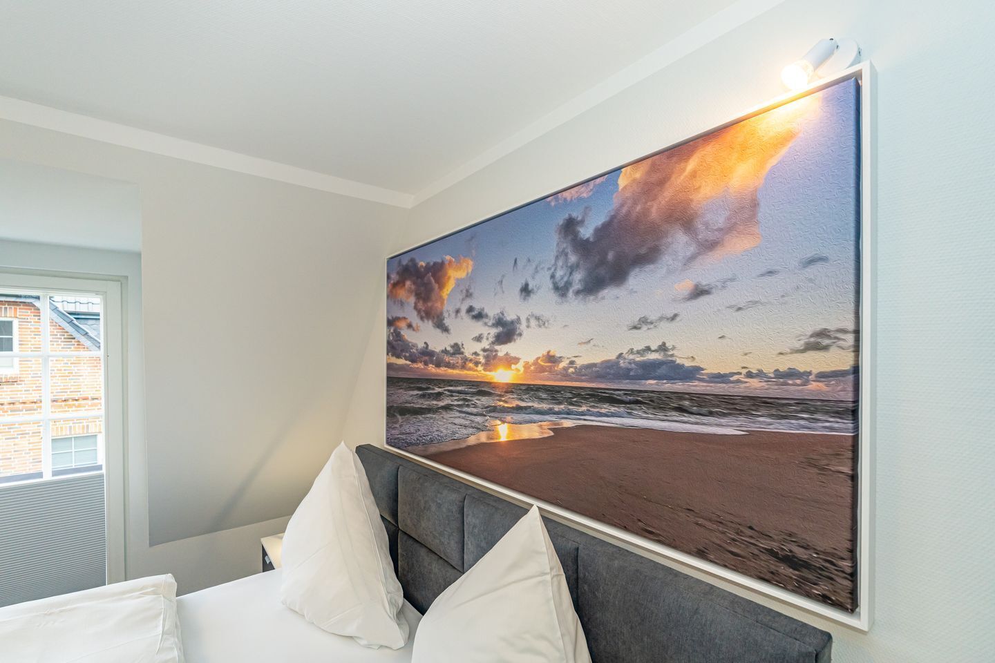 Een slaapkamer met een groot schilderij van een zonsondergang boven de oceaan, een grijs gestoffeerd hoofdeinde en witte beddengoed.