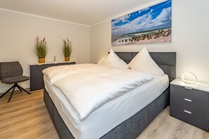 A modern bedroom with a large bed, bedside tables, a chair, and a painting of a beach scene.