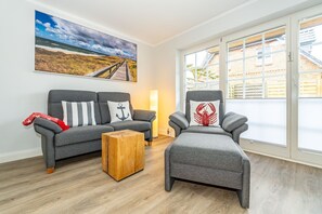 A living room with a grey sofa, a wooden coffee table, and a grey armchair with a lobster pillow. A large painting of a beach and sky is on the wall.