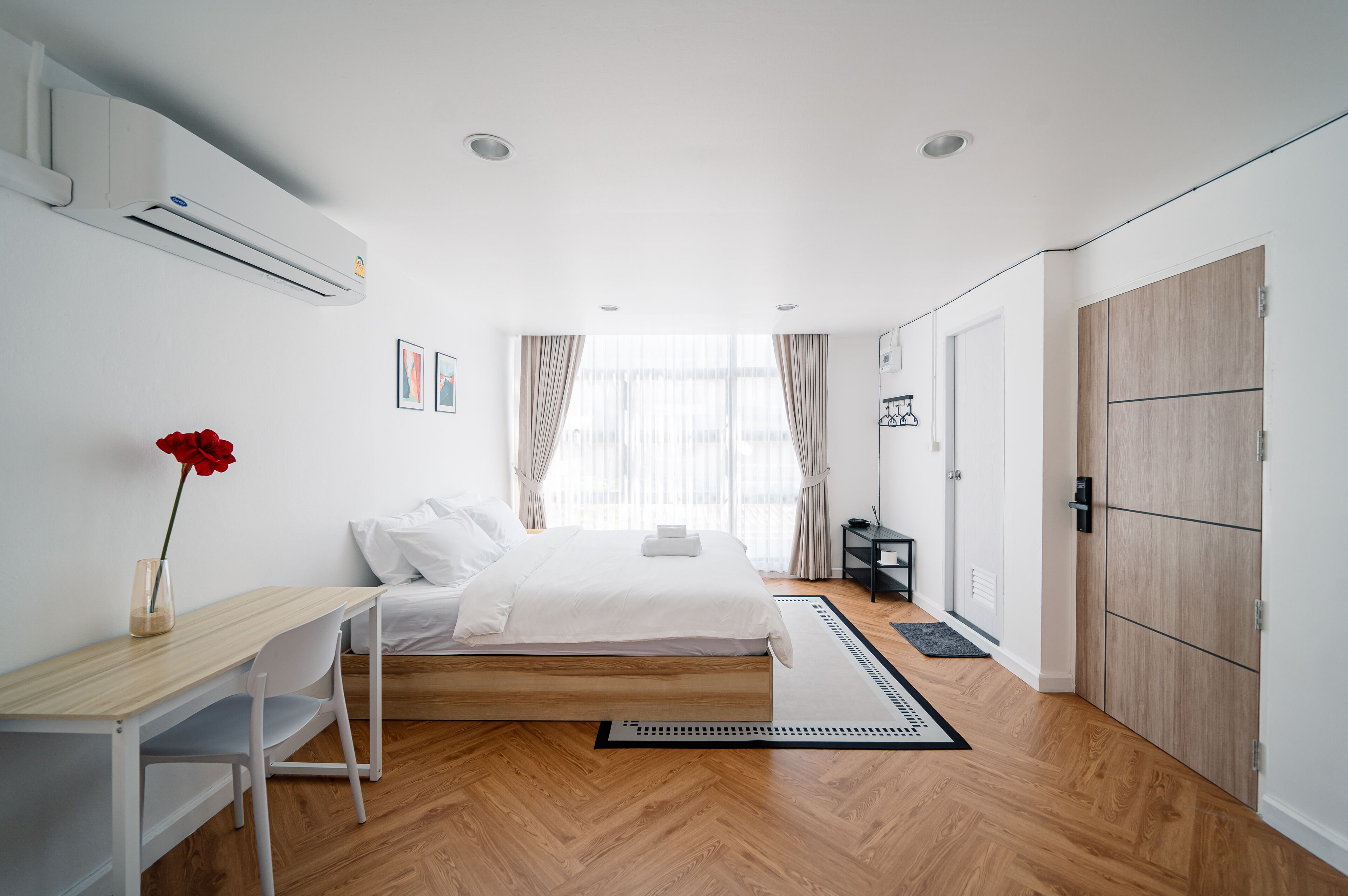 A modern hotel room with a wooden floor, a bed with white linens, a desk with a chair, a red flower vase, and a wardrobe.