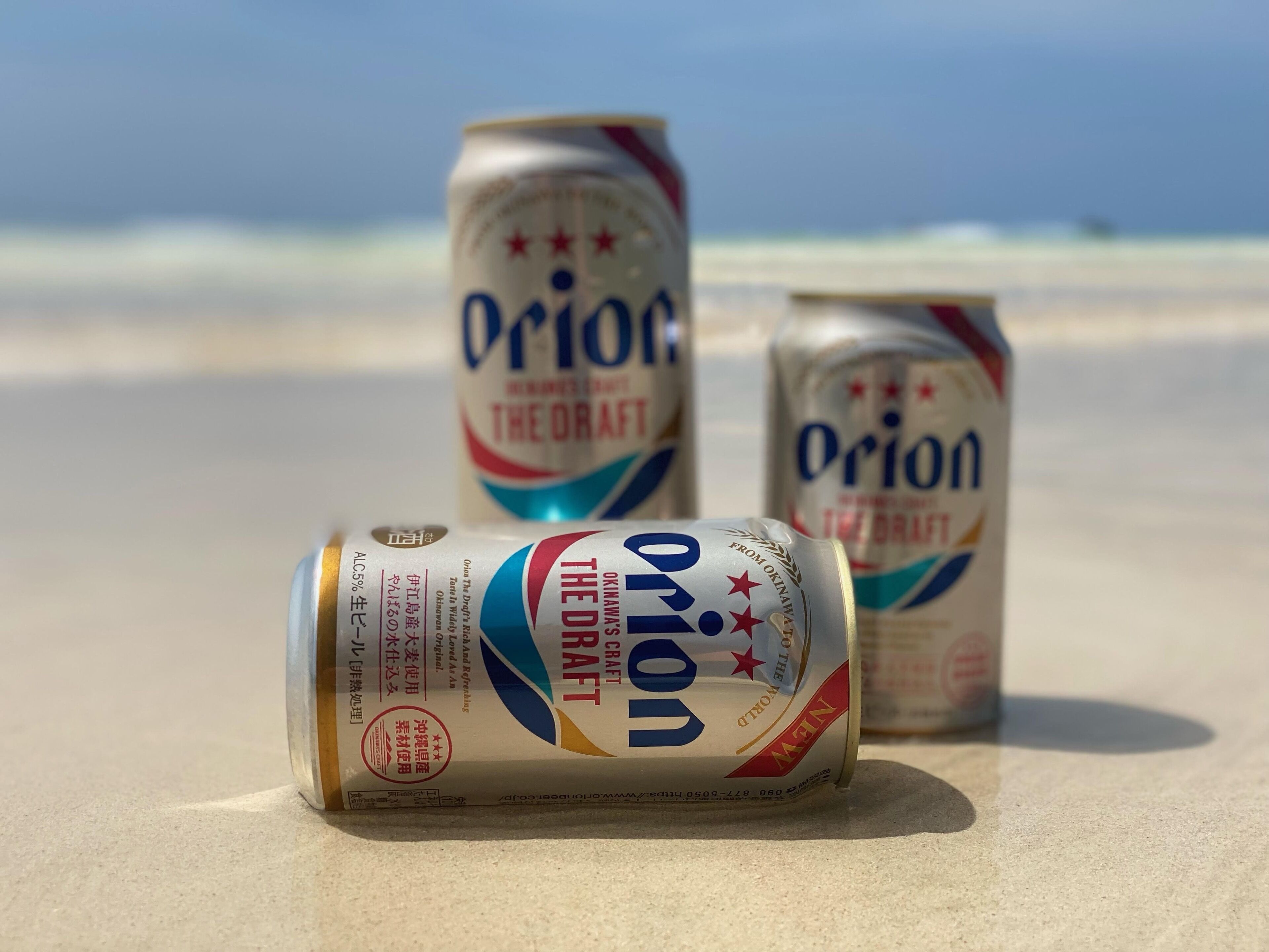 Three cans of Orion beer on a sandy beach with a clear sky in the background.