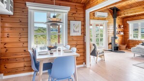 L’intérieur chaleureux d’un chalet avec des murs en bois, un coin repas avec des chaises bleues, une cheminée et une grande fenêtre donnant sur un paysage verdoyant.