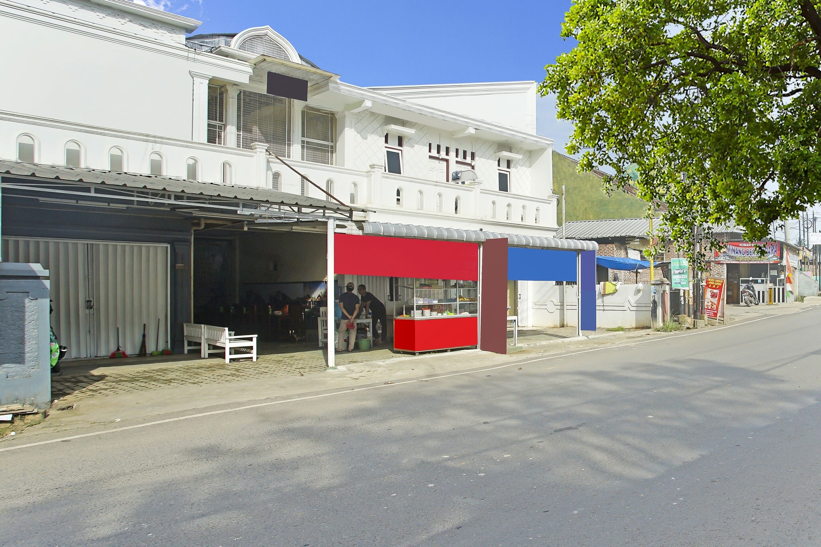 A street view with a white building, a red and blue kiosk, and a clear sky.