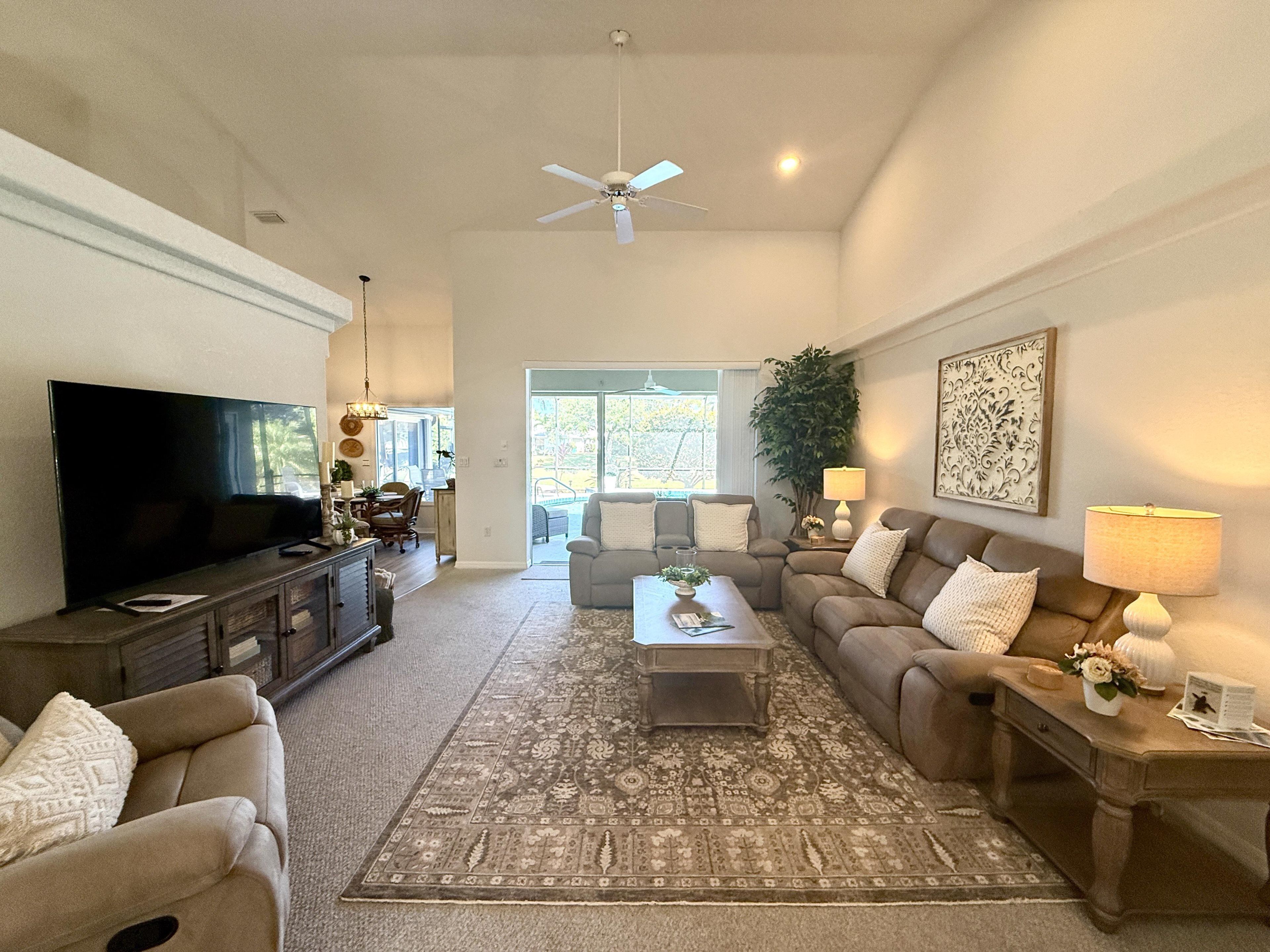 A spacious living room with a flat-screen TV, a ceiling fan, and a large window providing natural light.