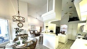 Une cuisine moderne avec des meubles blancs, un îlot central et un coin repas comprenant des chaises en osier et une table en verre.