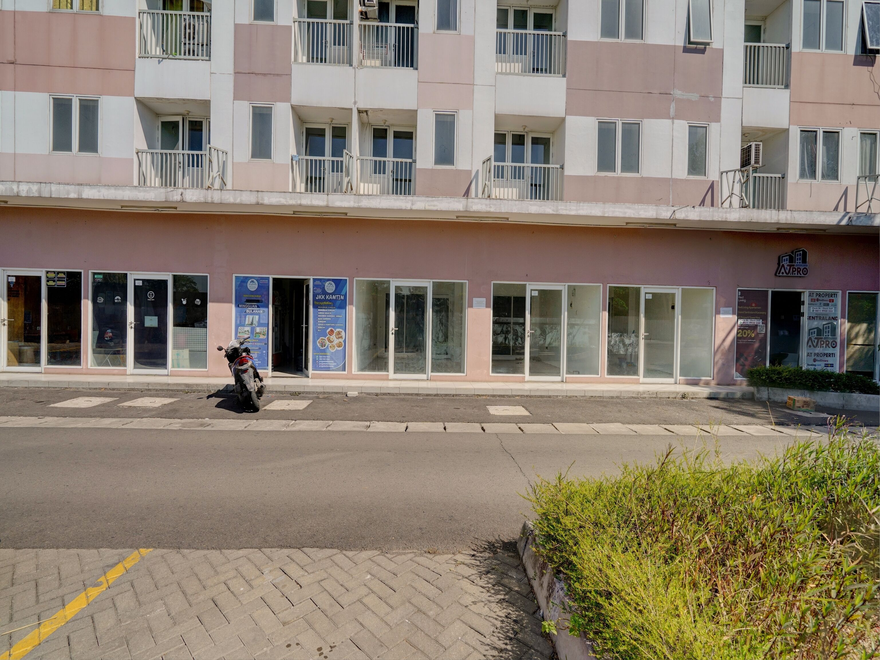 Un edificio de varios pisos con una fila de puertas y ventanas de vidrio, una motocicleta estacionada en la parte delantera, y un cartel anunciando servicios inmobiliarios.