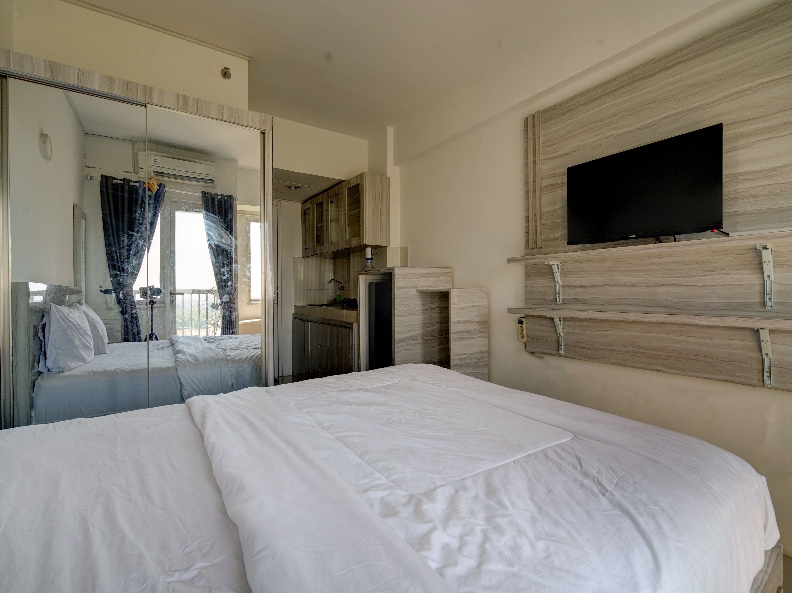 A hotel room with a large bed, a TV mounted on a wooden panel, a mirror, and a small kitchenette area.