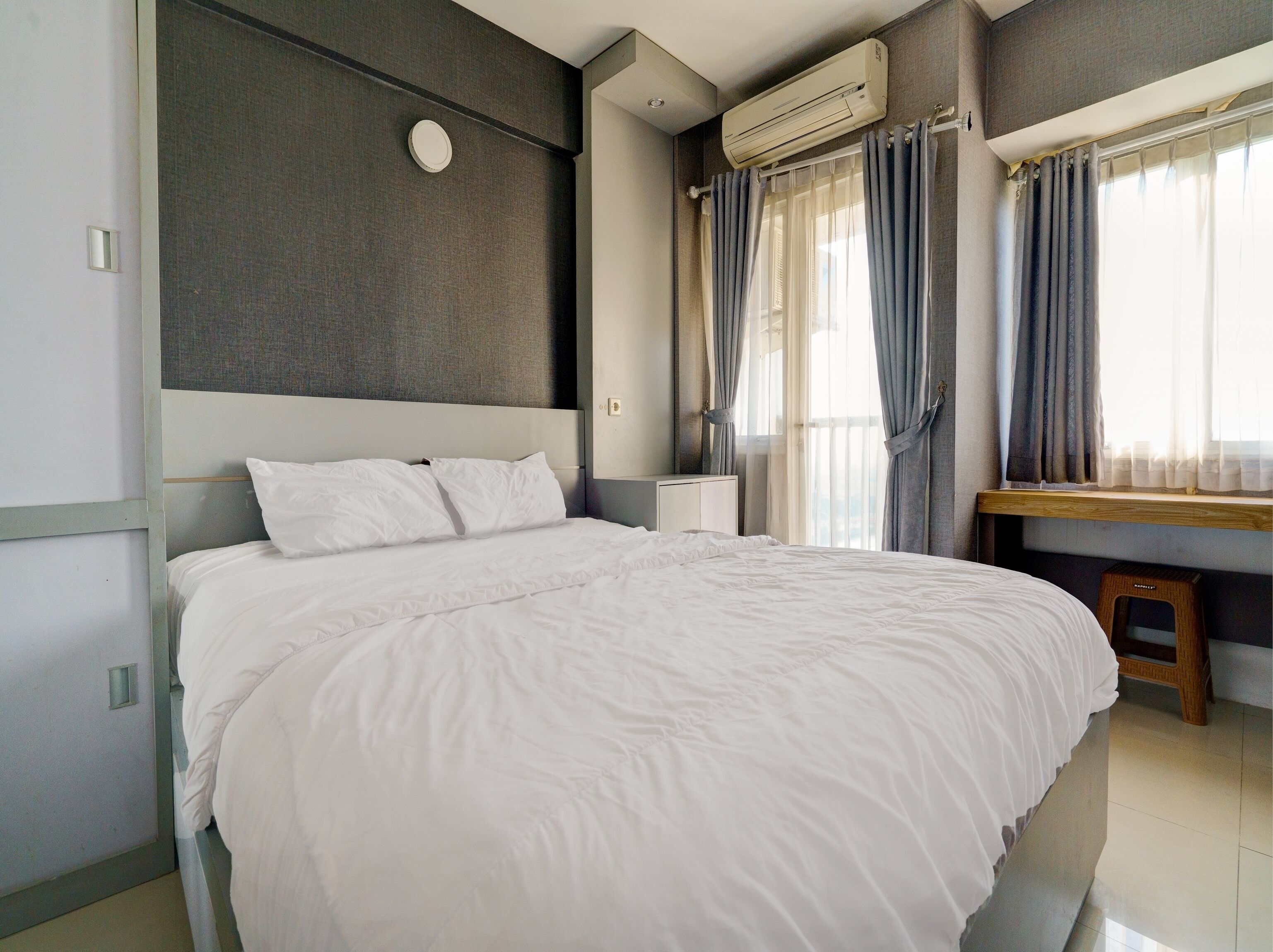 A neatly made bed in a hotel room with a large window, an air conditioning unit, and a small wooden stool.