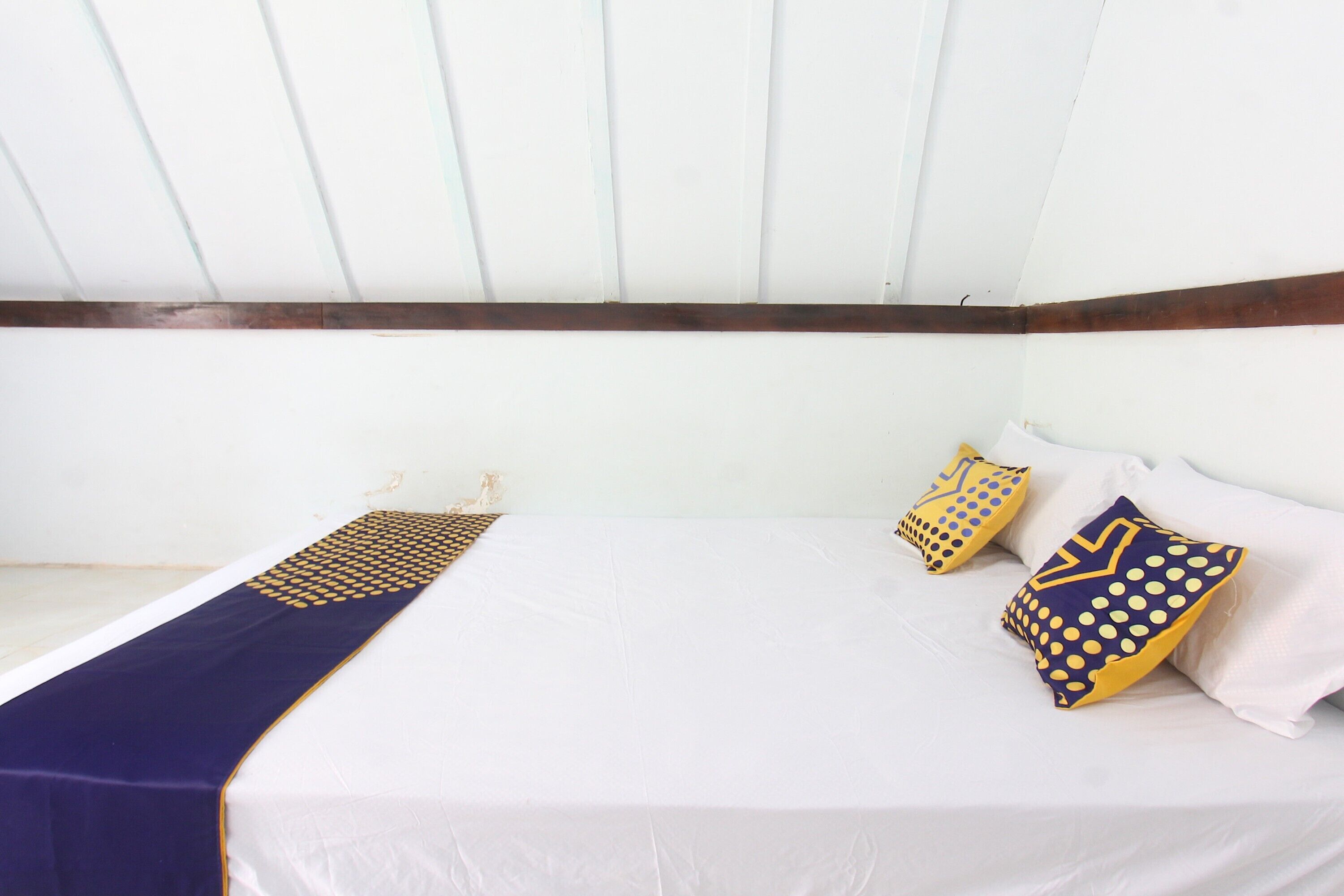A single bed with a white mattress, a dark blue bedspread with a gold pattern, and two decorative pillows with a yellow and blue polka dot design.