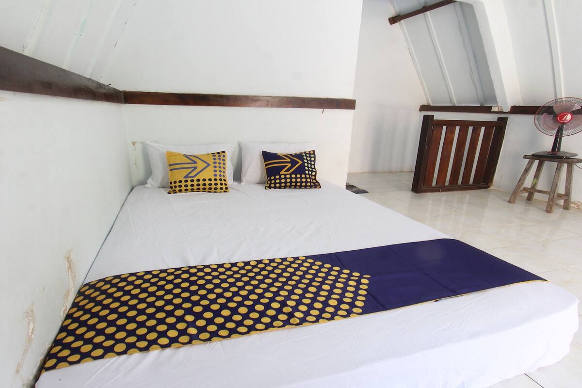 A bed with a white mattress, a blue and yellow polka dot blanket, and two patterned pillows. There is a wooden bench and a small fan in the room.