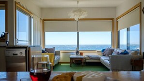 A modern living room with a large window overlooking the sea, a dining area with a table and chairs, and a kitchen area with a dishwasher and a sink. - AWESOME Beach House Bicheno (Glamorgan-Spring Bay)