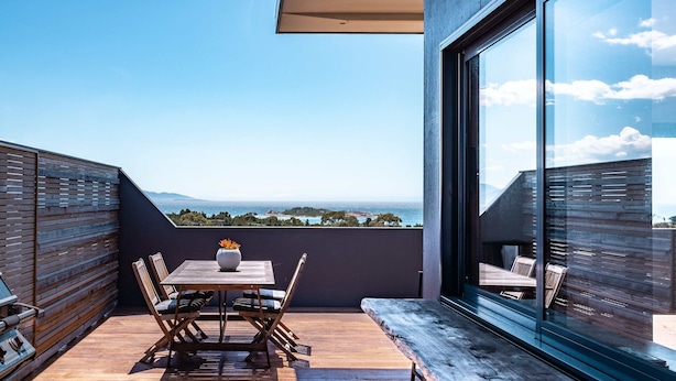 Uma varanda com mesa e cadeiras de madeira, um vaso com flores e vista para o mar e montanhas.