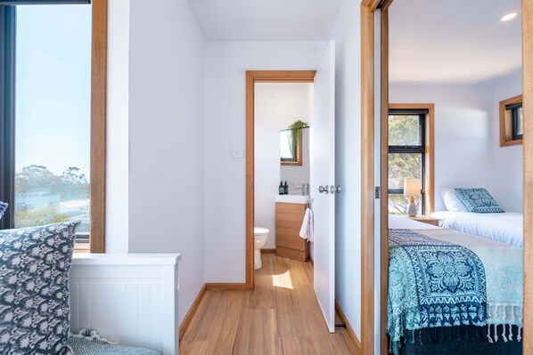 A modern hotel room with a wooden floor, a bed with a patterned bedspread, a bedside table with a lamp, and a view of the outdoors through a window. - Luxe PLUS! Incredible Views (Glamorgan-Spring Bay)