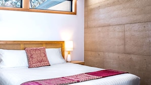 Quarto de hotel com uma cama grande, cabeceira de madeira, janela com vista para árvores, mesa de cabeceira com luminária e parede de destaque em concreto.