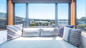 Quarto moderno com grandes janelas, área de estar confortável com almofadas e vista para a paisagem urbana.