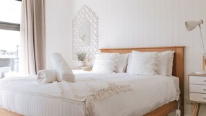 Ein ordentlich bezogenes Bett mit weißen Leinen und einem Holzkopfteil, eine Nachttischlampe und ein kleiner Nachttisch mit einer Lampe.