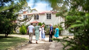 A group of people standing in a garden area in front of a house with a red roof and white walls.