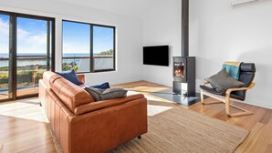 A modern living room with a brown leather sofa, a wooden floor, a large window with a view, a black TV, and a wood-burning stove. - Lucy (Glamorgan-Spring Bay)