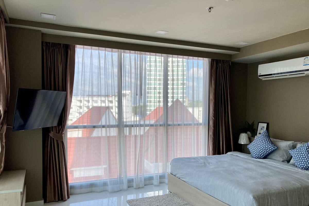 Quarto moderno com cama grande, TV de tela plana na parede e janela ampla com cortinas leves.