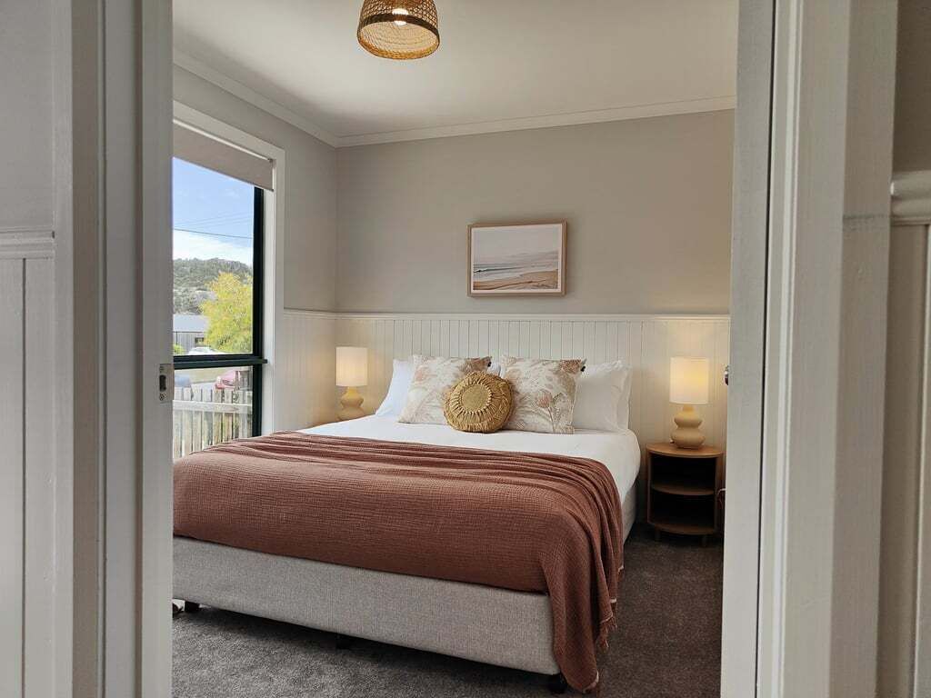 A bedroom with a large bed, two bedside tables with lamps, a framed picture on the wall, and a view of the outdoors through a window.