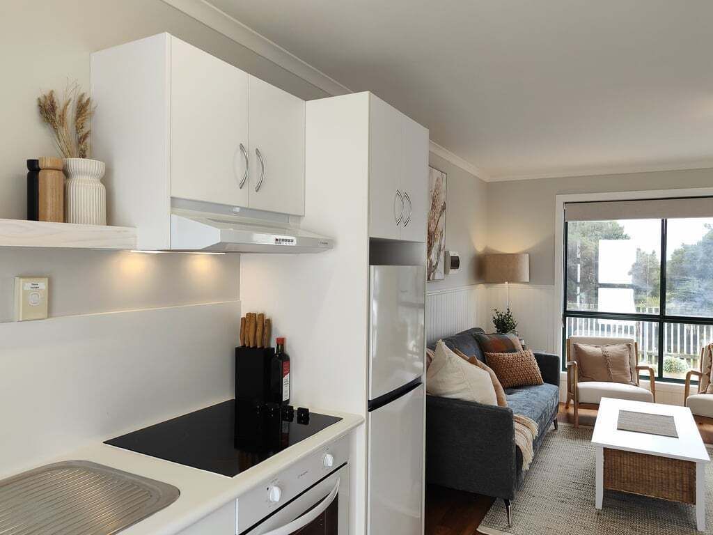 A modern kitchen with white cabinetry, a black induction cooktop, and a built-in oven. There is a dining area with a white table and chairs, and a large window providing a view of the outdoors.