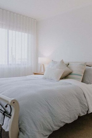A bedroom with a bed, pillows, a bedside table, and a window with sheer curtains.