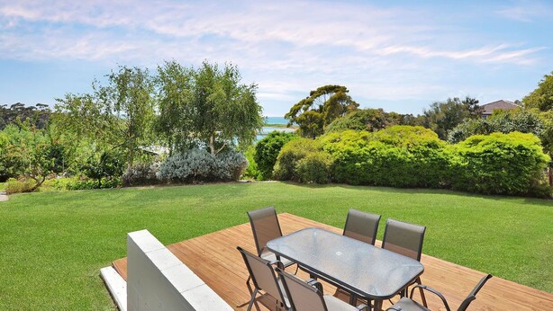 Une terrasse avec une table et des chaises, entourée d’une pelouse bien entretenue et de divers arbustes.