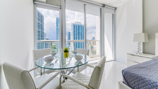 A modern hotel room with a balcony, a glass dining table, white furniture, and a bed with a quilted cover. - Apt 2505 · Brickell Icon STUDIO (Miami)