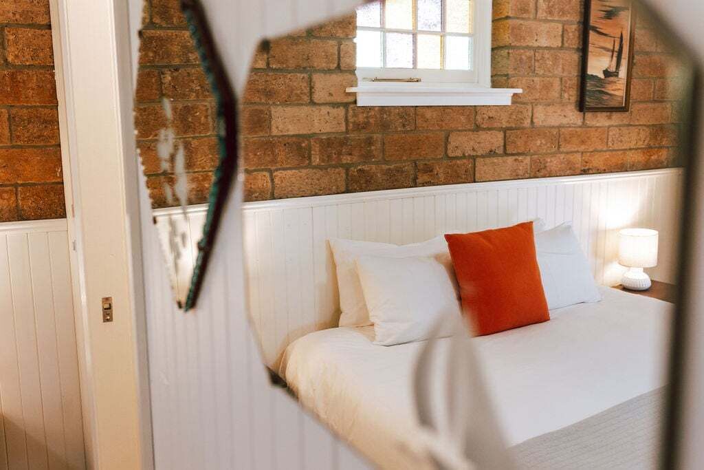 A bedroom with a brick wall, a window, a bed with white bedding and an orange pillow, and a bedside lamp.