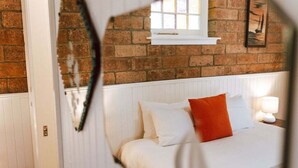 Une chambre avec un mur en briques, une fenêtre, un lit avec une literie blanche et un oreiller orange, et une lampe de chevet.