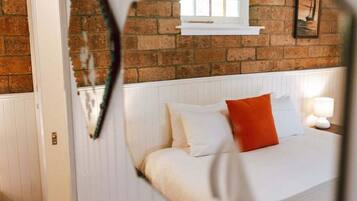 A bedroom with a brick wall, a window, a bed with white bedding and an orange pillow, and a bedside lamp.