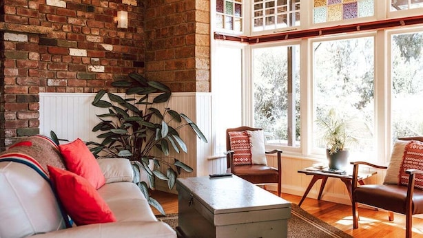 Un salon avec un mur en briques, un canapé blanc agrémenté de coussins rouges, une grande plante et une vue dégagée sur l’extérieur grâce à une grande fenêtre.