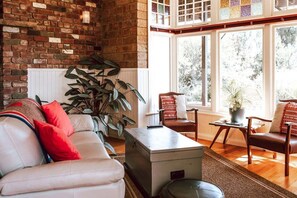 A living room with a brick wall, a white sofa with red cushions, a large plant, and a view through a large window.