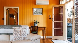 A room with a wooden paneled wall, a white sofa with a patterned pillow, a small table with a plant, and a door leading to an outdoor area with a vehicle. - The most beautiful guest retreat (Break O'Day)