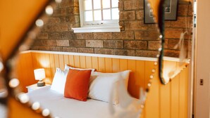 A bedroom with a brick wall, a window, a bed with white and orange pillows, and a nightstand with a lamp. - The most beautiful guest retreat (Break O'Day)