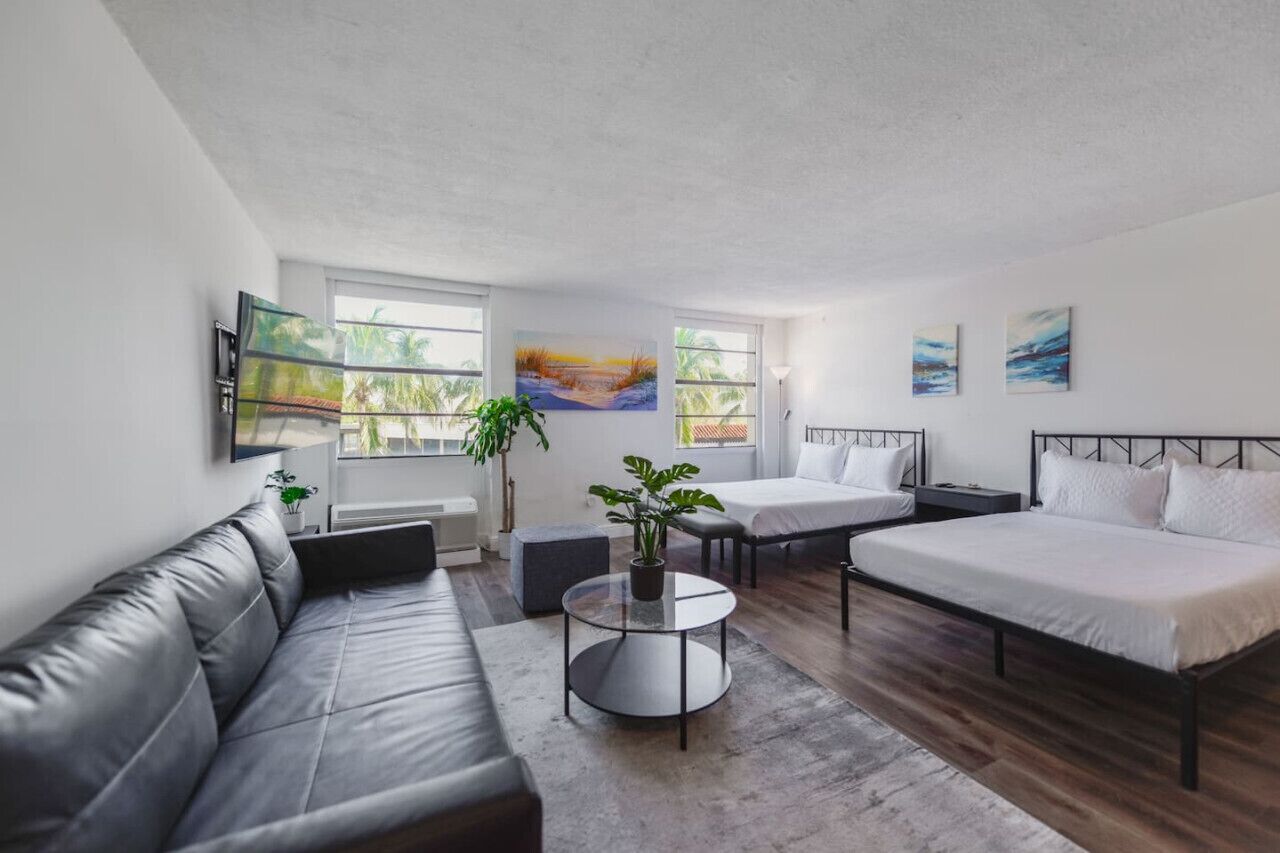 Quarto moderno e bem iluminado com um sofá de couro cinza, uma mesa de centro redonda de vidro, uma cama com roupa de cama branca e duas pinturas na parede.