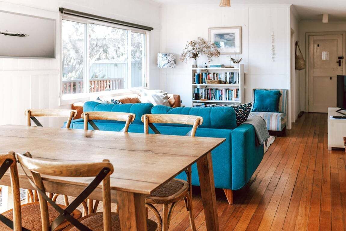 A living room with a blue sofa, wooden dining table, and chairs, a bookshelf, and a window with a view.