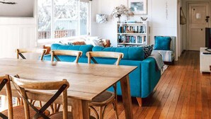 A living room with a blue sofa, wooden dining table, and chairs, a bookshelf, and a window with a view. - sloop cottage | beachfront | vibes (Break O'Day)