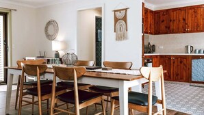 A dining area with a wooden table and chairs, a hanging macrame wall decoration, and a kitchen with wooden cabinets. - KAL's House - The place to be! (Glamorgan-Spring Bay)