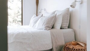 Un lit avec une literie blanche, une table d’appoint en osier et une lampe fixée au mur.